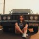 A woman relaxes in front of a vintage car under the warm sun, capturing a nostalgic moment.