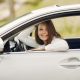 Cheerful female driver in white shirt sitting inside modern automobile and looking at camera through opened side window with smile during car trip in summer day