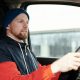 A bearded man wearing a hoodie and knitted cap driving a car, focused and attentive.